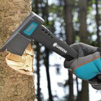 Bild einer Person, die eine kleine Axt mit dem Brand Gardena verwendet, um durch ein Baumstamm zu schlagen. Die Person trägt einen blauen Handschuh und der Baum hat rauhes Rindenwerk und wird von anderen Bäumen umgeben.